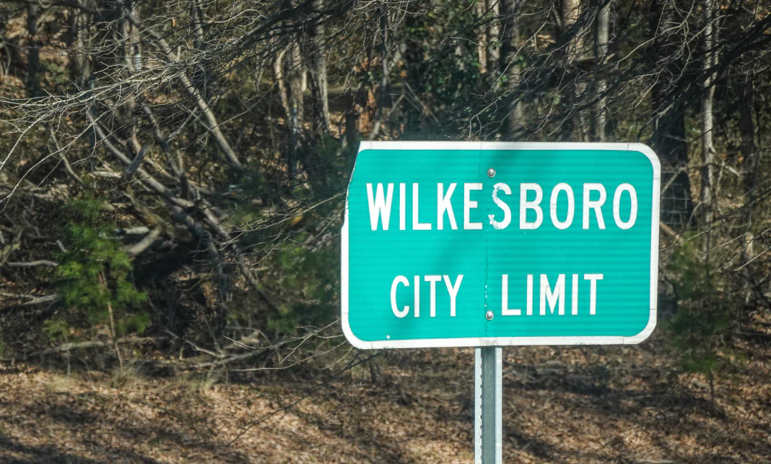 Desktop_Contact_Us_Plan_Your_Visit Green road sign reads Wilkesboro City Limit with a wooded area in the background.