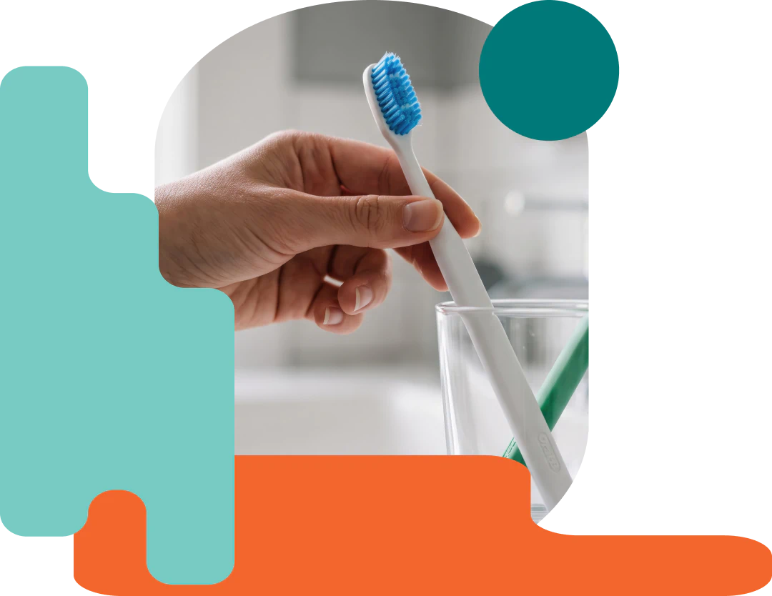 A hand places a white toothbrush with blue bristles into a clear glass, with another green toothbrush visible inside. Abstract teal and orange shapes accent the image.