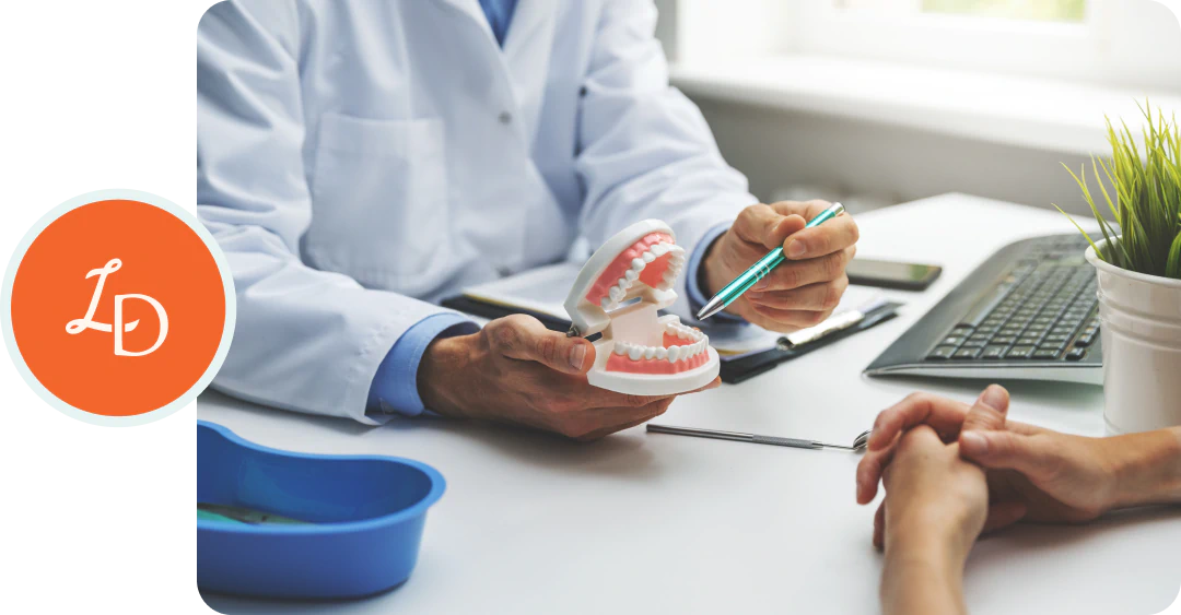 A dentist in a white coat uses a pen to explain dental care with a plastic tooth model to a patient sitting across the desk, near a tray and a computer keyboard.